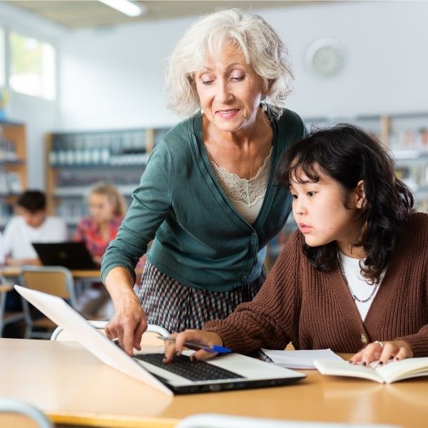 Teacher assisting a student with writing on a laptop in a classroom, surrounded by books and other students, representing personalized support and structured writing instruction with the Jane Schaffer Writing Program®.