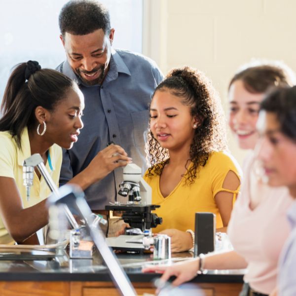 Science teacher guiding a diverse group of students using a microscope in a classroom, representing hands-on STEM learning and the integration of writing skills across disciplines.
