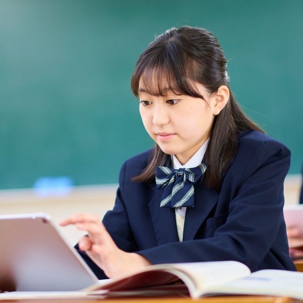 Student in school uniform using a tablet in a classroom setting, with an open book in front and a chalkboard in the background, representing technology integration in education.